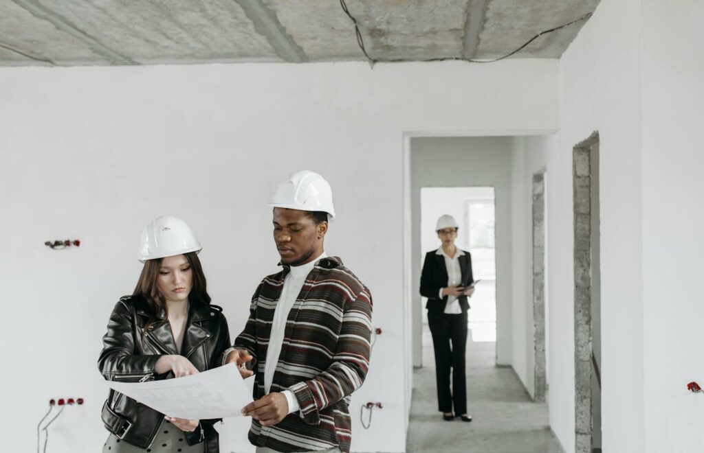 A couple wearing hard hats examines building plans inside an unfinished property with a realtor observing.