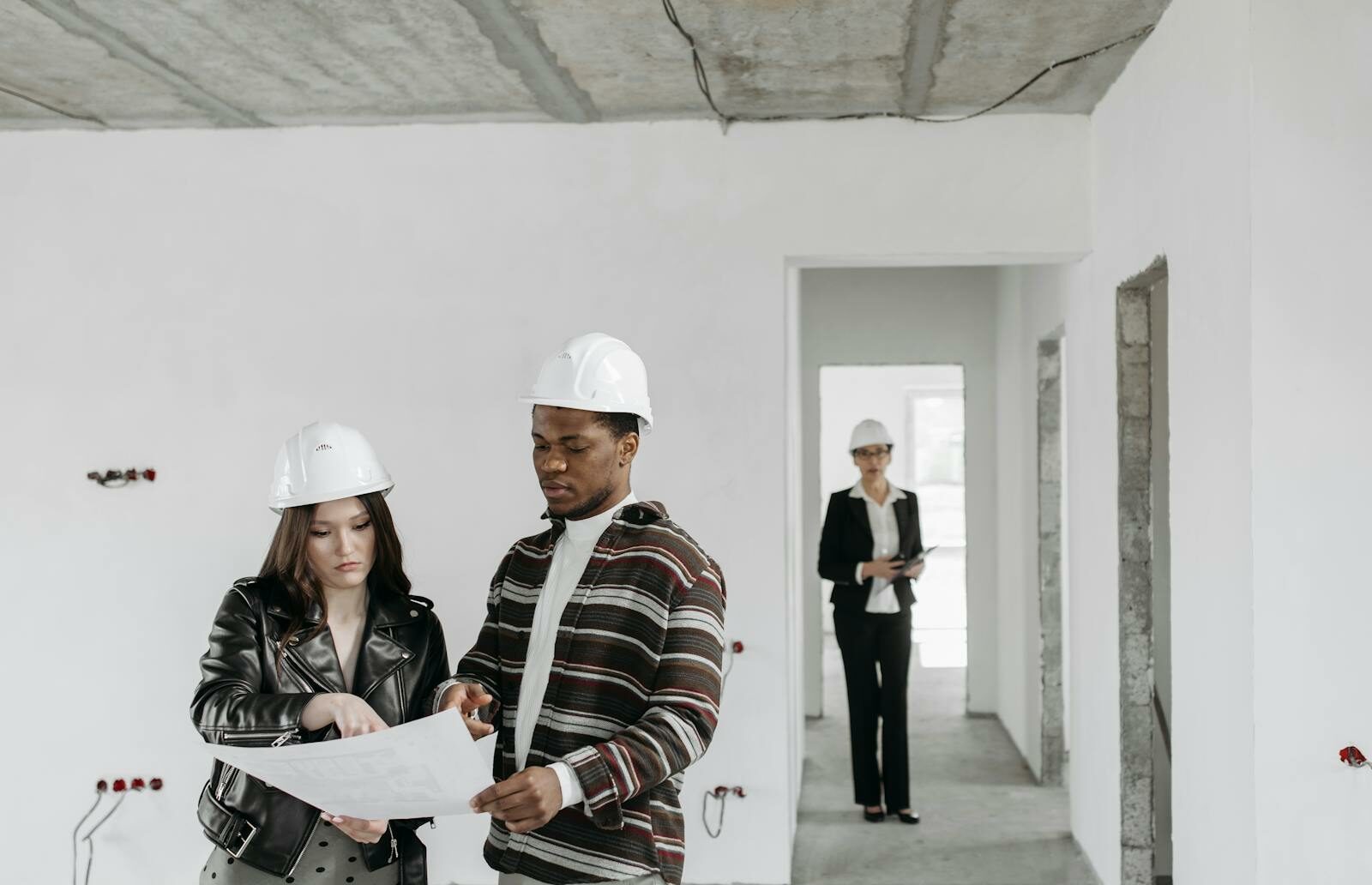 A couple wearing hard hats examines building plans inside an unfinished property with a realtor observing.